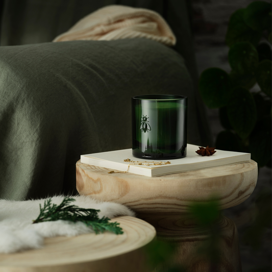 Grüne Duftkerze in einem Glas mit geprägtem Bienensymbol, dekorativ auf einem Holztablett mit Buch und Schmuck arrangiert, im Hintergrund ein Sofa mit grüner Decke.