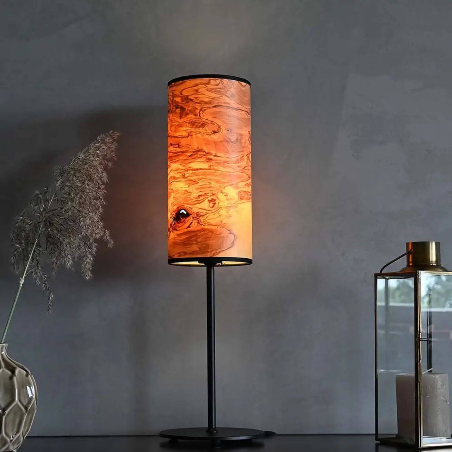 Tischlampe mit zylindrischem Holzfurnier-Lampenschirm auf schwarzem Metallfuß, eingeschaltet, auf einem Sideboard neben Dekoration.
