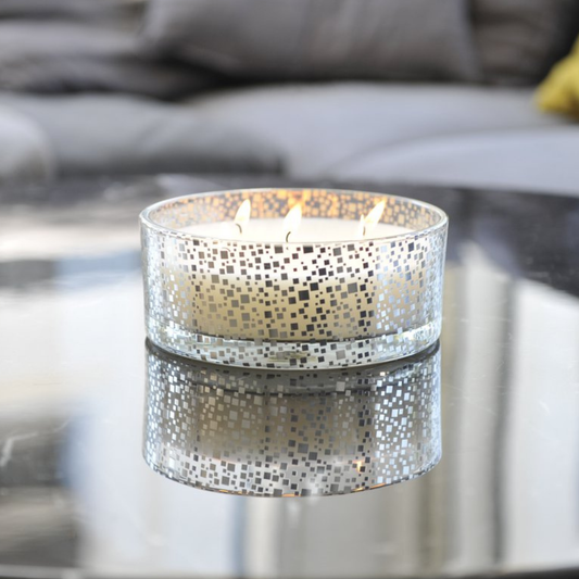 Aulica Vanille-Kerze in silbernem Mosaikglas mit drei brennenden Dochten auf Glascouchtisch, Spiegelung im Tisch sichtbar, graues Sofa im Hintergrund