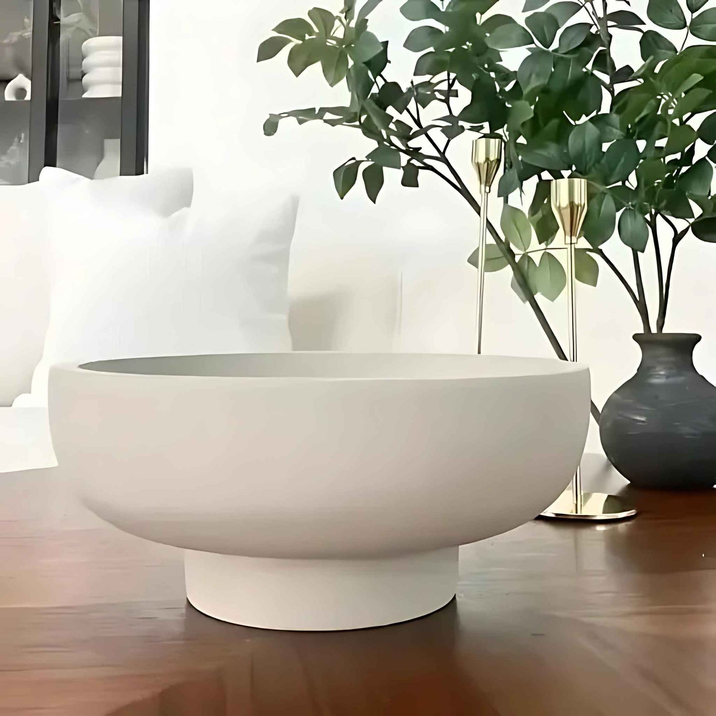 Große, skulpturale Betonschale in Beige auf einem Holztisch, dekoriert mit goldfarbenen Kerzenhaltern und einer grünen Vase im Hintergrund.