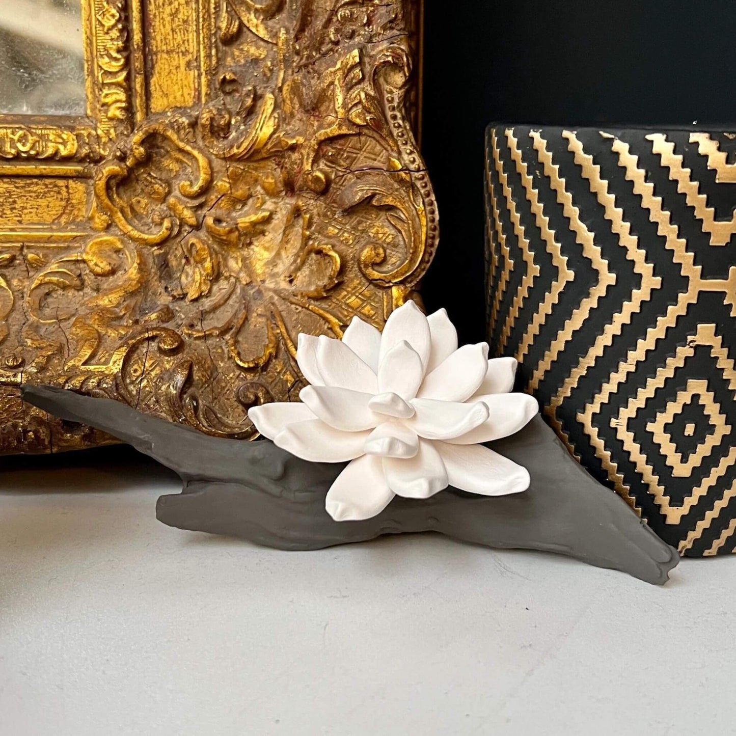 Weiße Keramikblume auf Treibholzdeko, platziert vor einem goldenen Spiegelrahmen und schwarzer Vase.