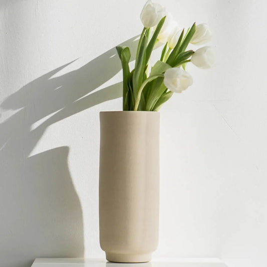 Hohe Steingutvase mit weißen Tulpen, aufgestellt vor einer weißen Wand.