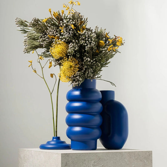 Blaue Loop Vase mit runden Ringen, gefüllt mit gelben Blüten, auf hellem Podest