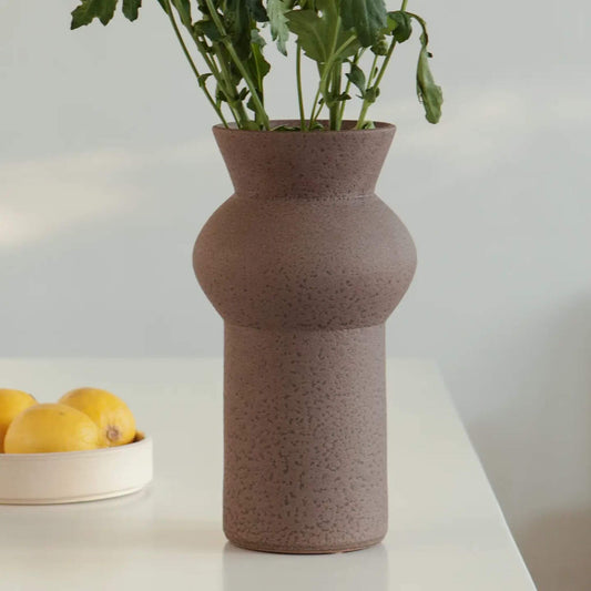 Braune Vase mit bauchiger Mitte, darin grüne Stiele von frischen Blumen.