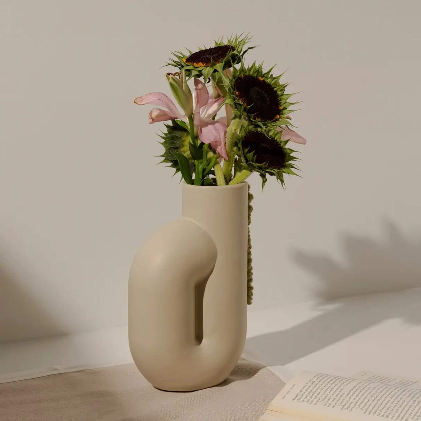 Cremefarbene Vase mit Blumen auf hellem Podest, vor neutralem Hintergrund.