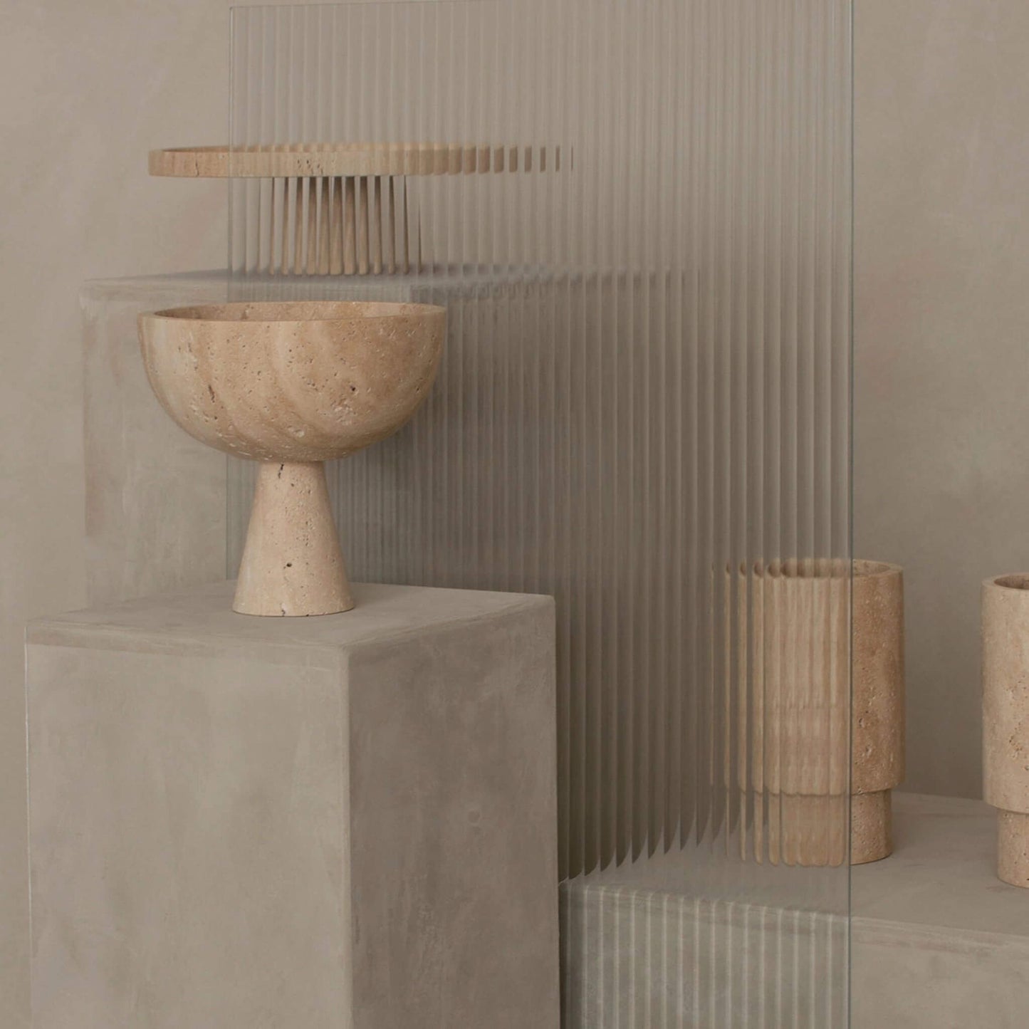 Podestschale aus beigem Travertinstein auf kubischem Sockel, minimalistisches Interieur mit Glaswand im Hintergrund.