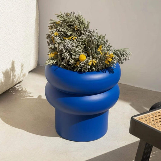 Blauer Blumentopf mit runder Wellenform, bepflanzt mit grünen Zweigen und gelben Blüten.