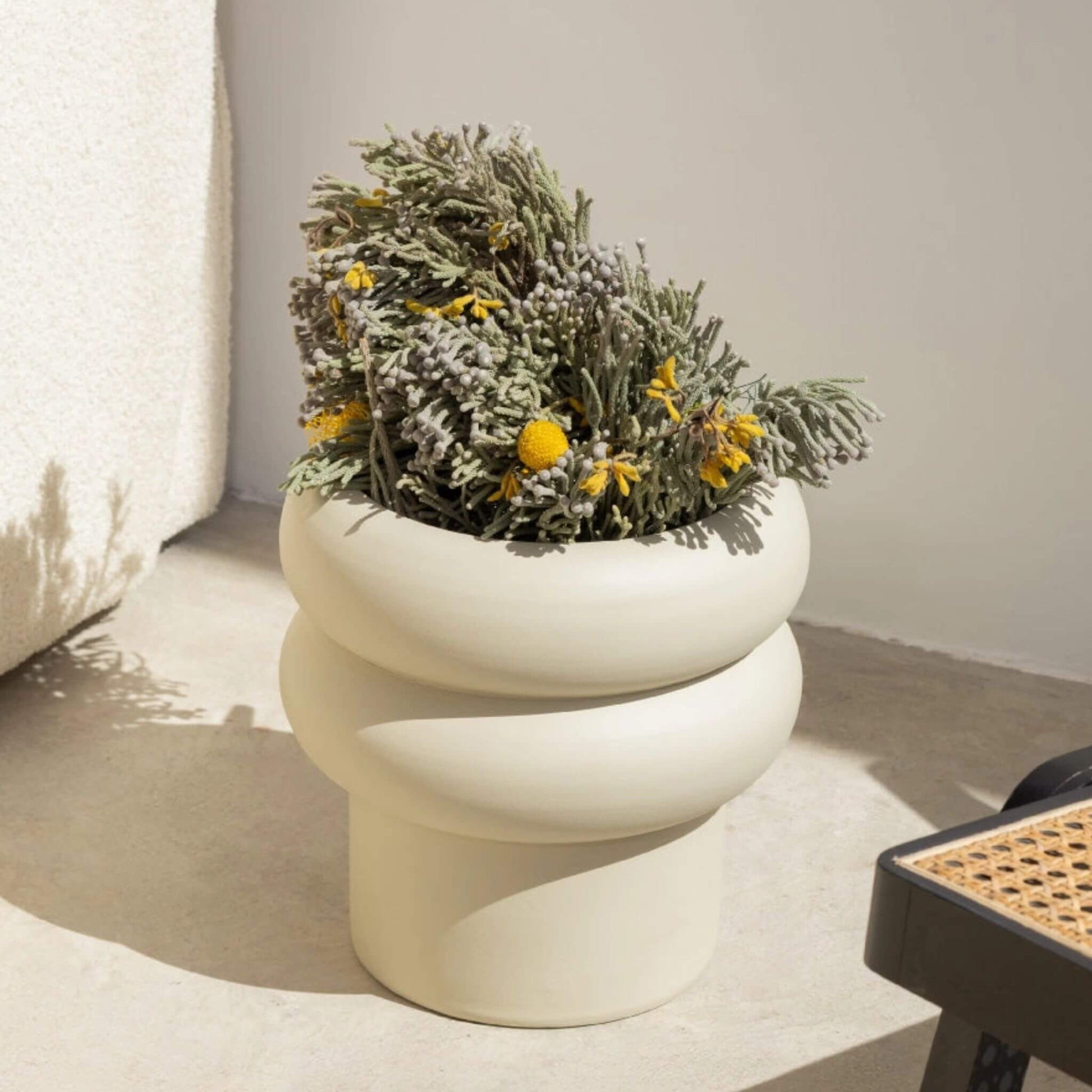 Off-white Blumentopf mit runder Wellenform, bepflanzt mit grünen Zweigen und gelben Blüten.