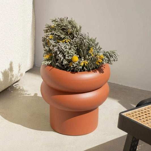 Terrakotta-Blumentopf mit runder Wellenform, bepflanzt mit grünen Zweigen und gelben Blüten.