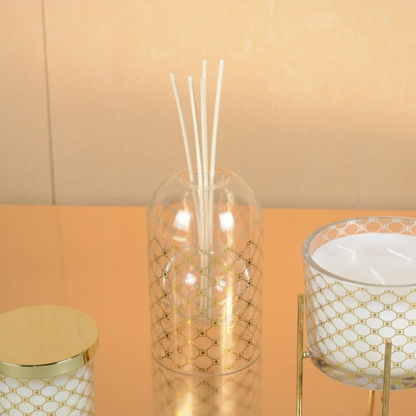 Aulica Moschus-Diffusor mit Wabenmuster-Glas zwischen passender Duftkerze und Kerzenhalter auf Spiegelfläche, goldorange Hintergrund