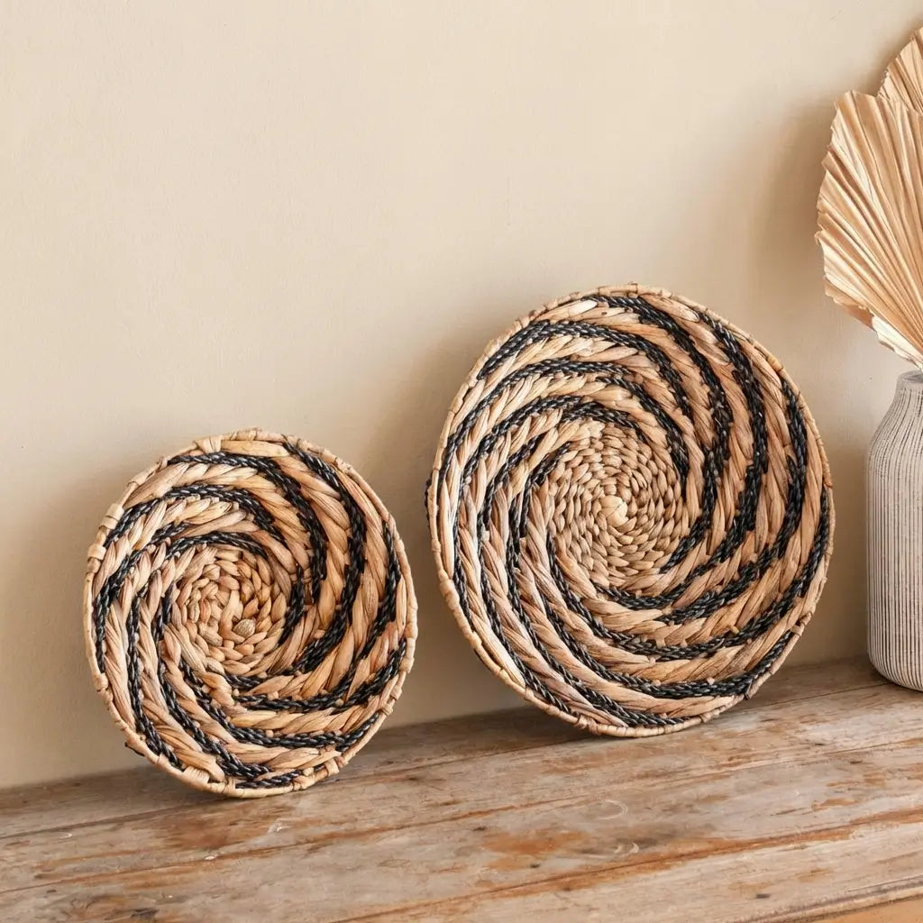 Rundes Wanddekor Bandung aus Wasserhyazinthe mit schwarzem Spiral-Muster im Boho-Stil, dekorativ auf einem Holzsideboard platziert.