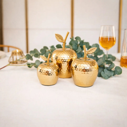 Drei goldene Deko-Äpfel auf Tisch mit Eukalyptus und Glas.