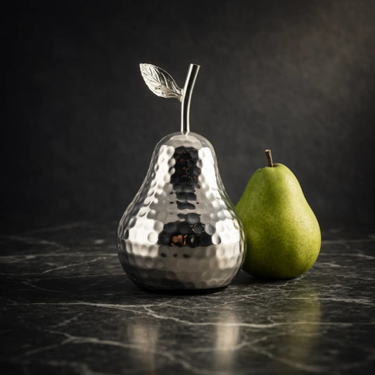 Glänzende Deko-Birne aus Aluminium auf dunklem Marmortisch neben frischer grüner Birne in elegantem, stilvollem Ambiente.