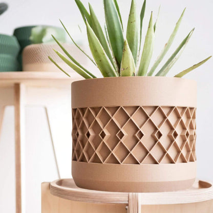Großes beiges Pflanzgefäß mit geometrischem Muster und Aloe, auf einem hellen Holzständer neben weiteren Pflanzgefäßen.