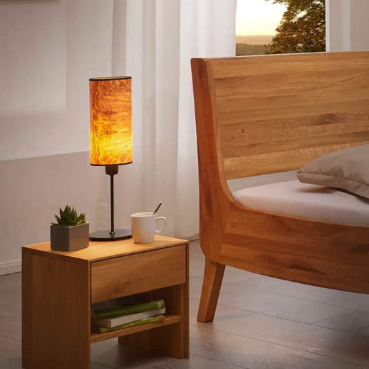 Nachttischlampe mit Holzfurnier-Schirm leuchtet warm auf einem Holznachttisch neben einem Bett.