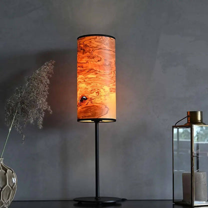 Tischlampe mit zylindrischem Holzfurnier-Lampenschirm auf schwarzem Metallfuß, eingeschaltet, auf einem Sideboard neben Dekoration.