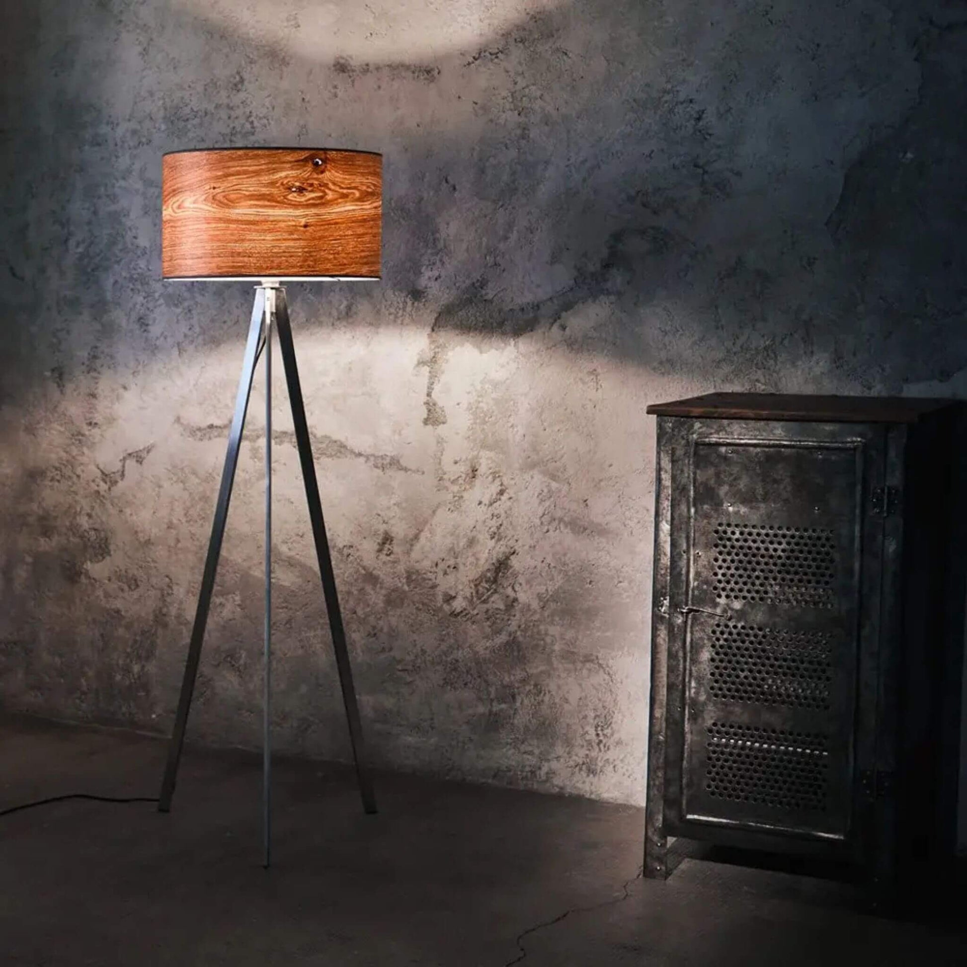 Dreibeinige Stehlampe mit rundem Lampenschirm aus Eichenholzfurnier, strahlt warmes Licht in einem industriellen Raum mit Betonwand und Metallschrank.