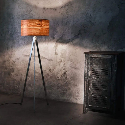 Dreibeinige Stehlampe mit rundem Lampenschirm aus Eichenholzfurnier, strahlt warmes Licht in einem industriellen Raum mit Betonwand und Metallschrank.