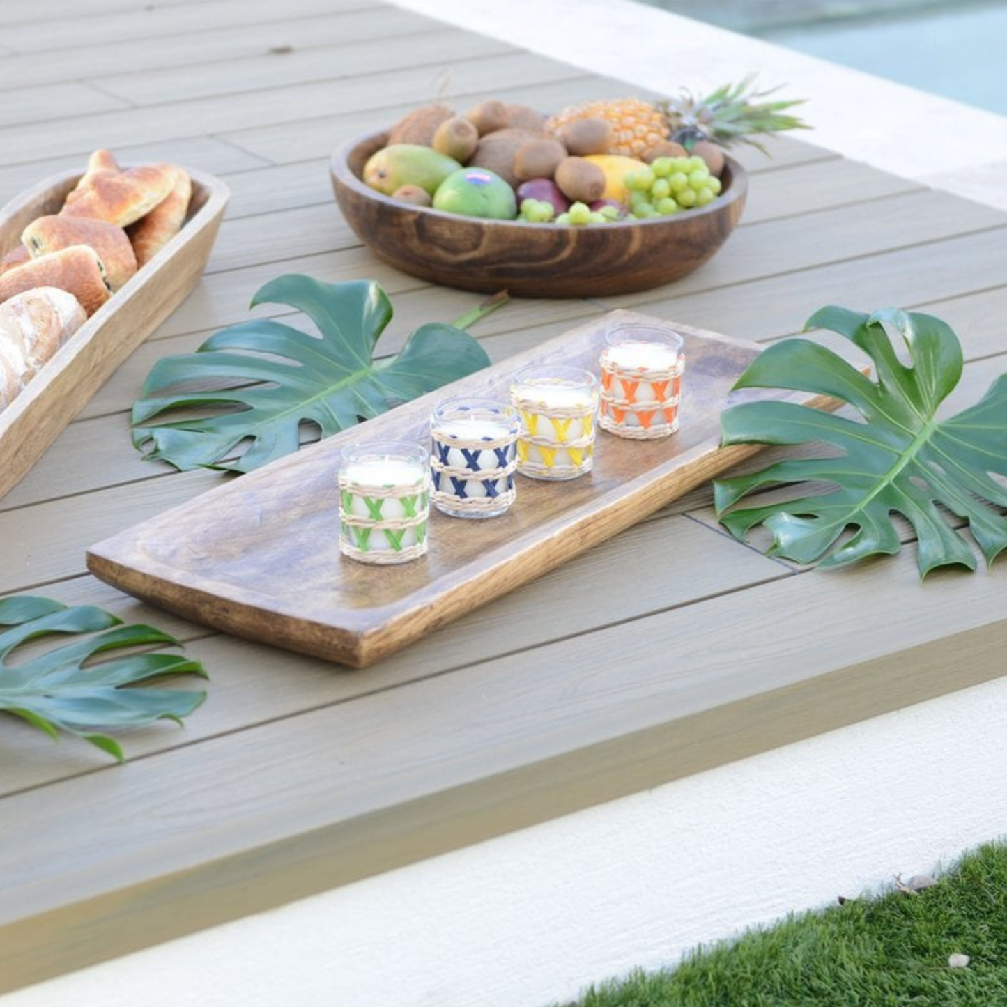 Vier Aulica Salbei-Duftkerzen mit Rattangeflecht in Grün, Blau, Gelb und Orange auf Holztablett, Terrassentisch mit Monstera-Blättern und Obstschale im Hintergrund