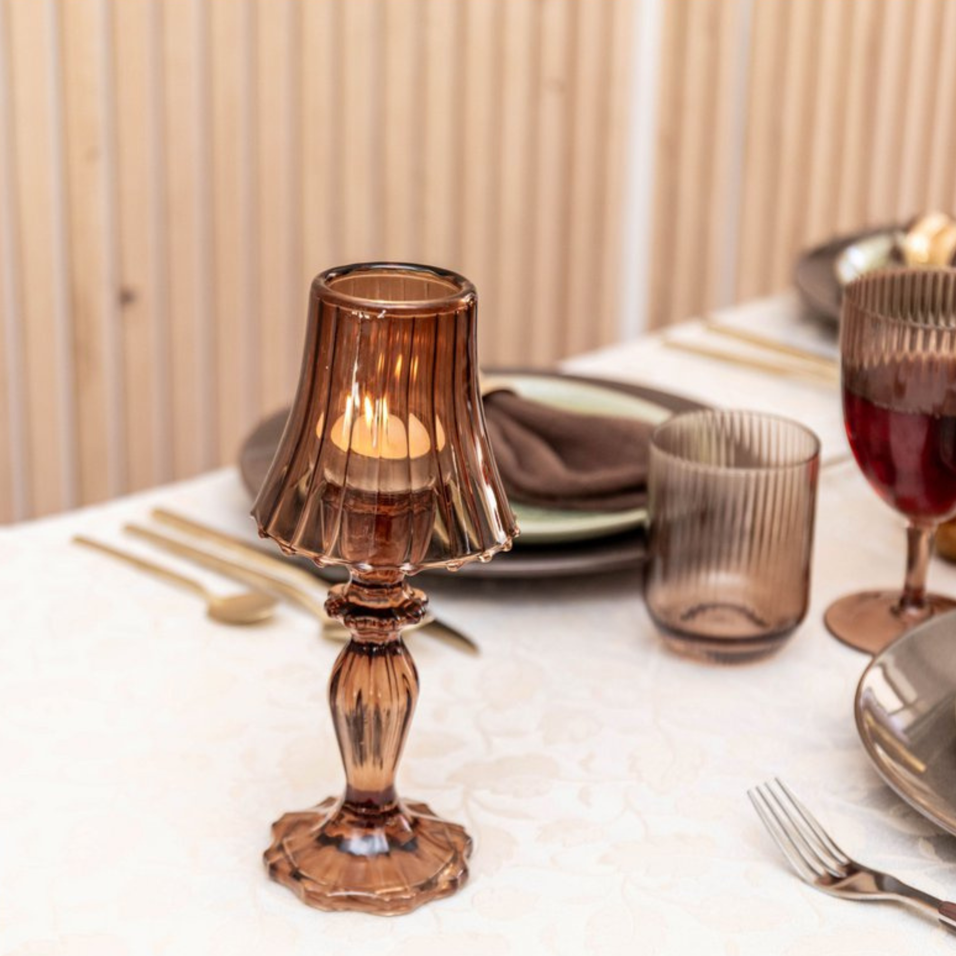 Aulica Kerzenhalter in Lampenform aus braunem Glas mit brennendem Teelicht auf gedecktem Esstisch, umgeben von Goldbesteck, braunen Gläsern und grünen Tellern