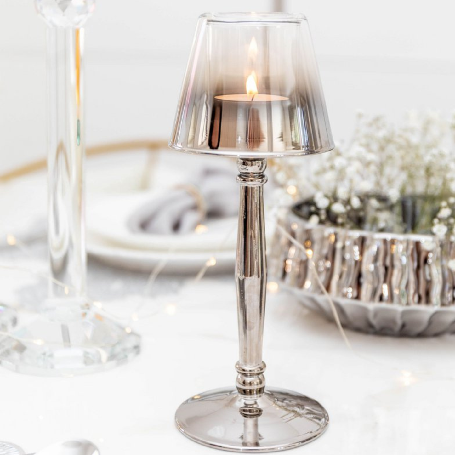 Aulica Kerzenhalter Silber mit brennendem Teelicht, warmes Licht durch Rauchglas-Schirm, weißer Tischhintergrund mit Dekoration