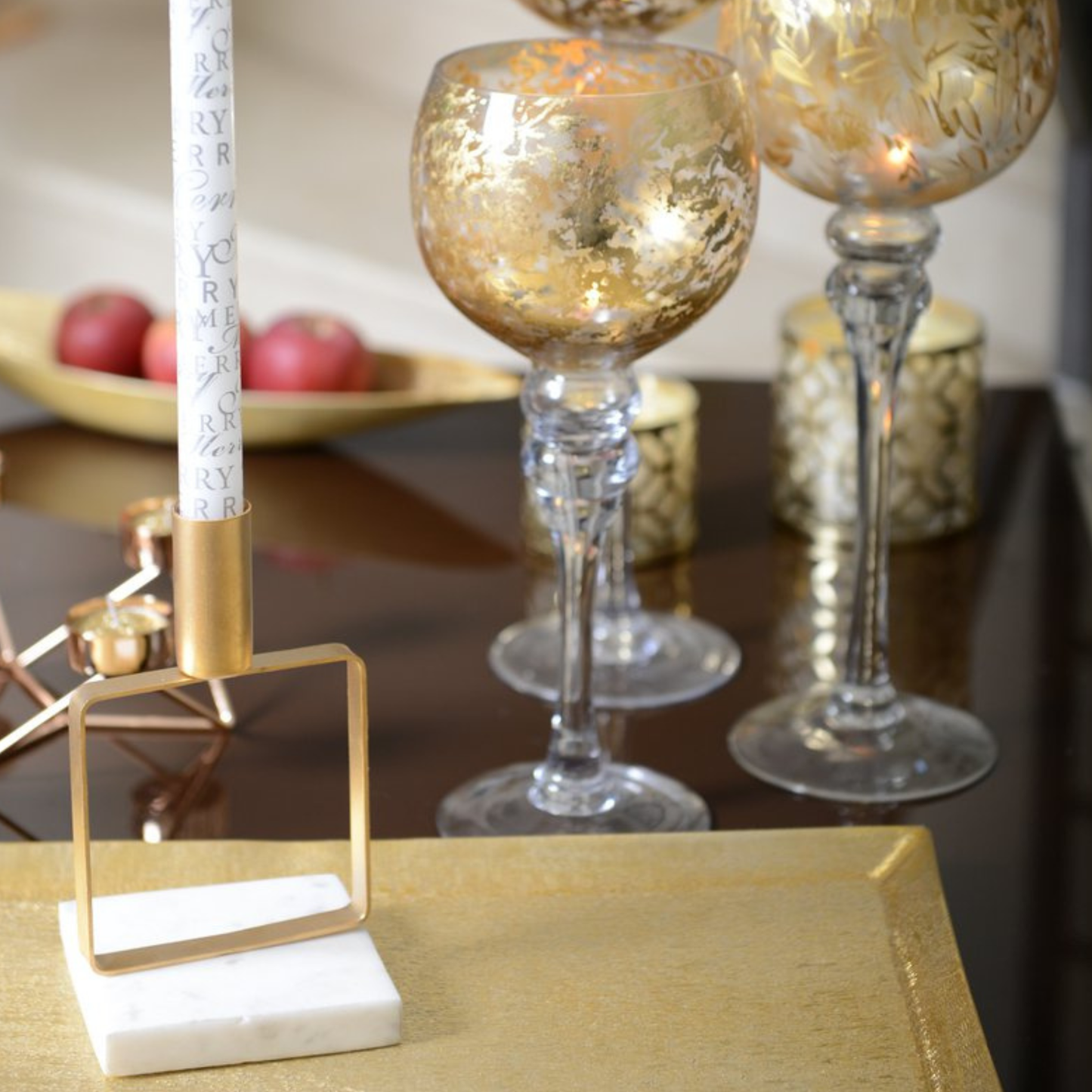 Nahaufnahme des Aulica Kerzenhalters mit weißem Marmorsockel und quadratischem Mattgold-Rahmen, bestückt mit weißer dekorierter Stabkerze, auf goldenem Tablett neben goldverzierten Weingläsern, festliche Tischatmosphäre.