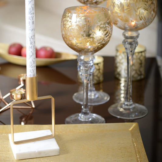 Nahaufnahme des Aulica Kerzenhalters mit weißem Marmorsockel und quadratischem Mattgold-Rahmen, bestückt mit weißer dekorierter Stabkerze, auf goldenem Tablett neben goldverzierten Weingläsern, festliche Tischatmosphäre.