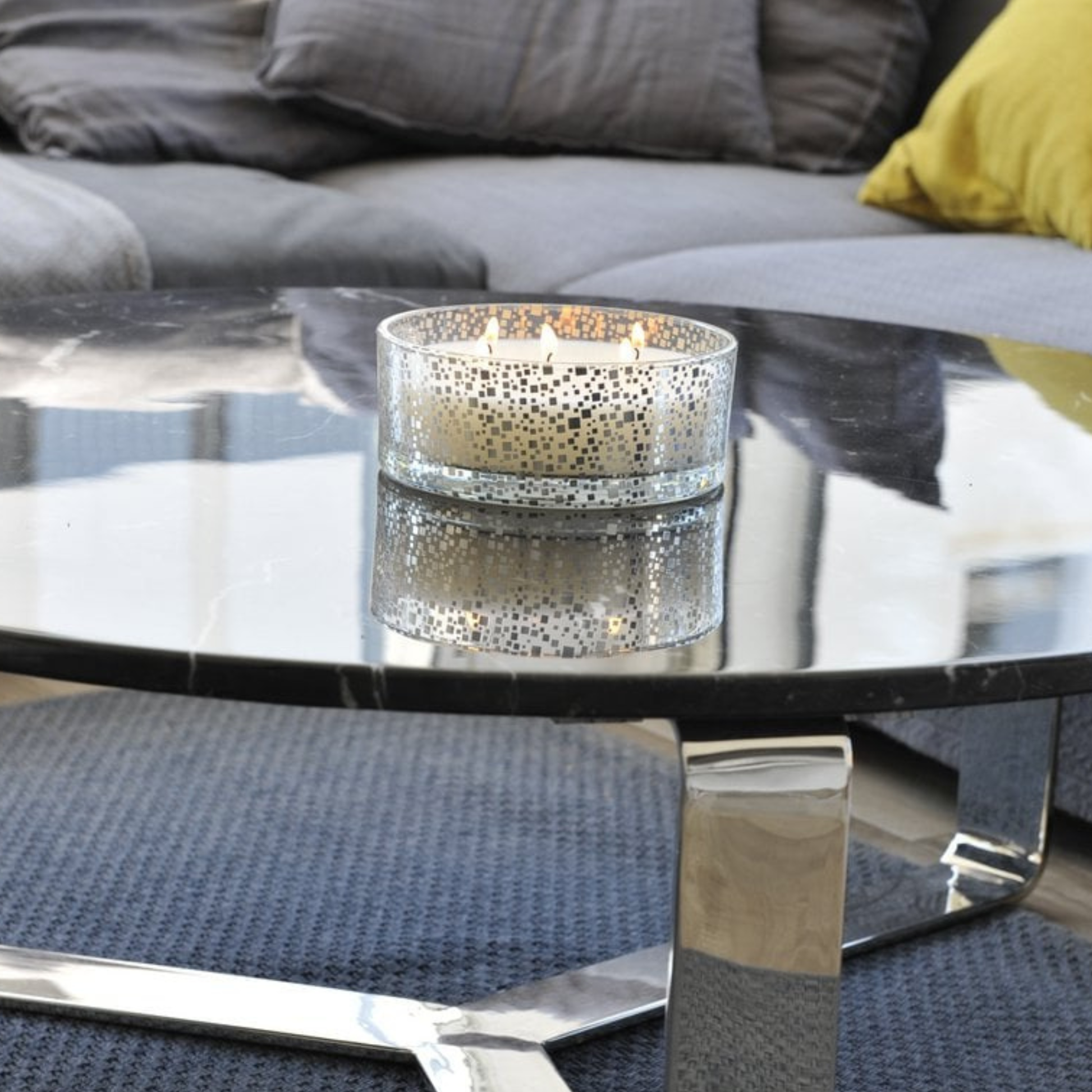 Aulica Vanille-Kerze in silbernem Mosaikglas mit drei brennenden Dochten auf Glascouchtisch, Spiegelung im Tisch sichtbar, graues Sofa im Hintergrund