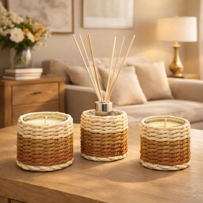 Aulica Moschus-Diffusor mit Geflecht-Mantel zwischen zwei Duftkerzen auf Holzcouchtisch, beiges Sofa und Blumenstrauß im Hintergrund