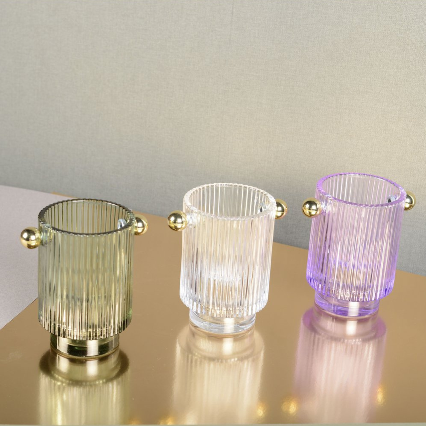 Drei Aulica Teelichthalter aus geripptem Glas in Grün, Klar und Lila mit goldenen Kugelgriffen und Standfuß auf goldfarbener Spiegelfläche