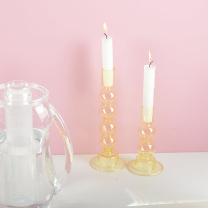Zwei gelbe Glas-Kerzenhalter von Aulica mit gestapelten Kugeln als Schaft, mit brennenden weißen Stabkerzen, auf weißer Oberfläche vor rosa Hintergrund, daneben ein transparenter Glaskrug