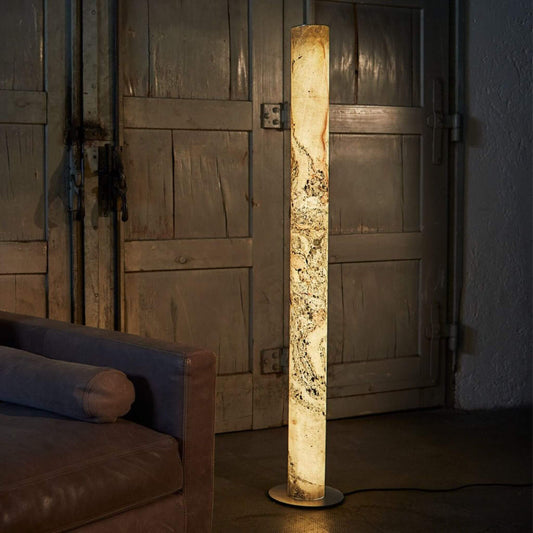 Stehlampe „Columna“ aus Stein in der Ausführung Weißperle, leuchtend neben einem Ledersofa vor rustikaler Holzwand.