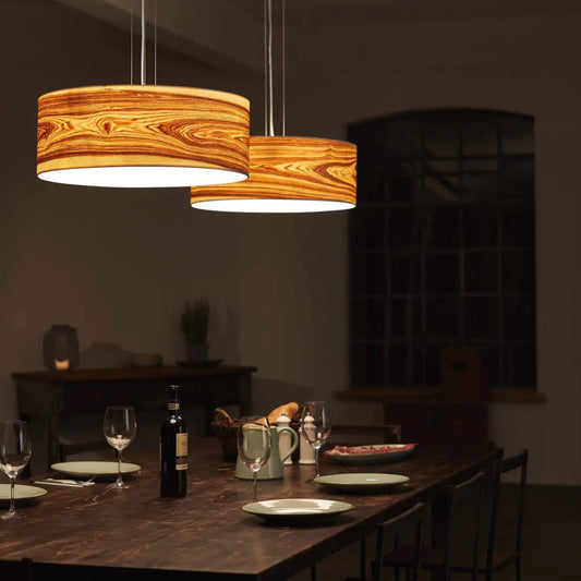 Pendelleuchte Discus 55 mit rundem Lampenschirm aus Oliveschenfurnier, über einem gedeckten Esstisch hängend, leuchtend im warmen Licht.