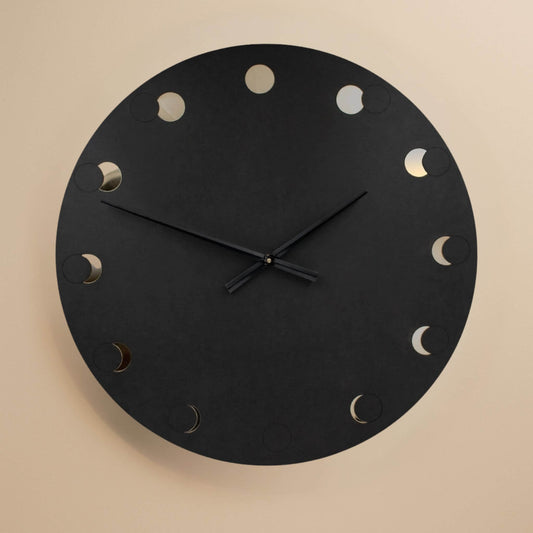 Freigestellte schwarze Wanduhr mit Mondphasen-Design und minimalistischen Zeigern auf beigem Hintergrund.