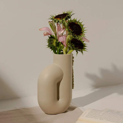 Cremefarbene Vase mit Blumen auf hellem Podest, vor neutralem Hintergrund.