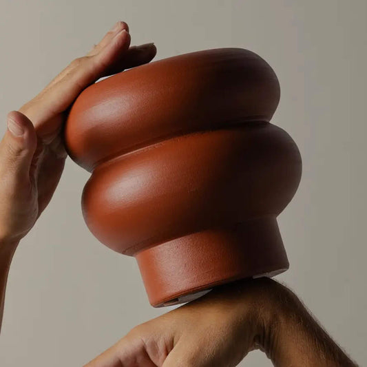 Hände halten kleine Terrakotta-Vase mit bauchiger Form gegen neutralen Hintergrund.