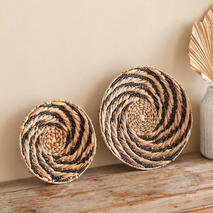 Rundes Wanddekor Bandung aus Wasserhyazinthe mit schwarzem Spiral-Muster im Boho-Stil, dekorativ auf einem Holzsideboard platziert.