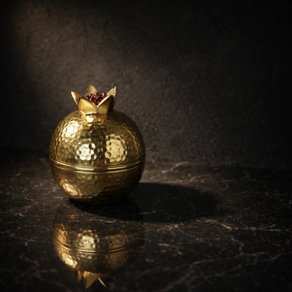 Goldene Granatapfel-Dose aus gehämmertem Aluminium auf schwarzem Marmor mit dunklem, strukturiertem Hintergrund und dramatischer Beleuchtung.
