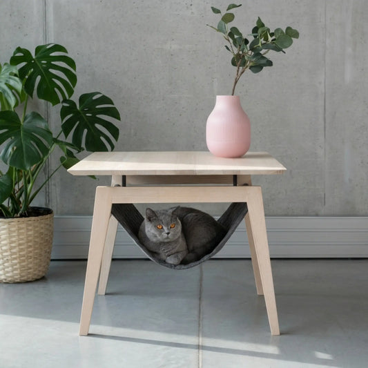 Graue Katze liegt entspannt in einem Katzenbett in Form einer Stoffhängematte unter modernen Tisch Kikko aus Eschenholz